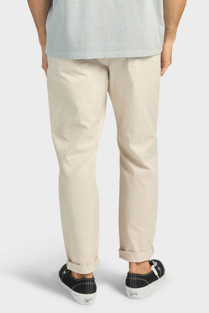 A person stands facing away, wearing light beige Academy Brand Oahu Pant, a light gray shirt, and black low-top sneakers with white soles, set against a plain light background.