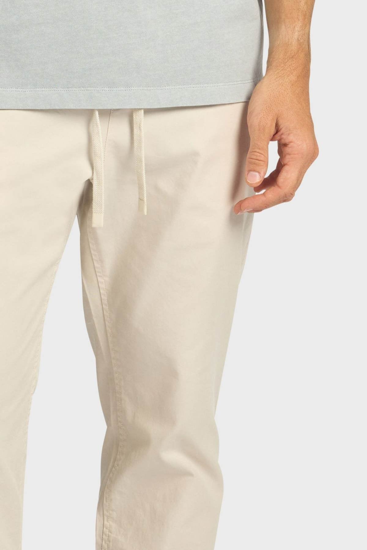 Wearing the Academy Brand Oahu Pant in light beige cotton twill, paired with a light gray shirt, a person stands with their right hand relaxed by their side against a plain background.