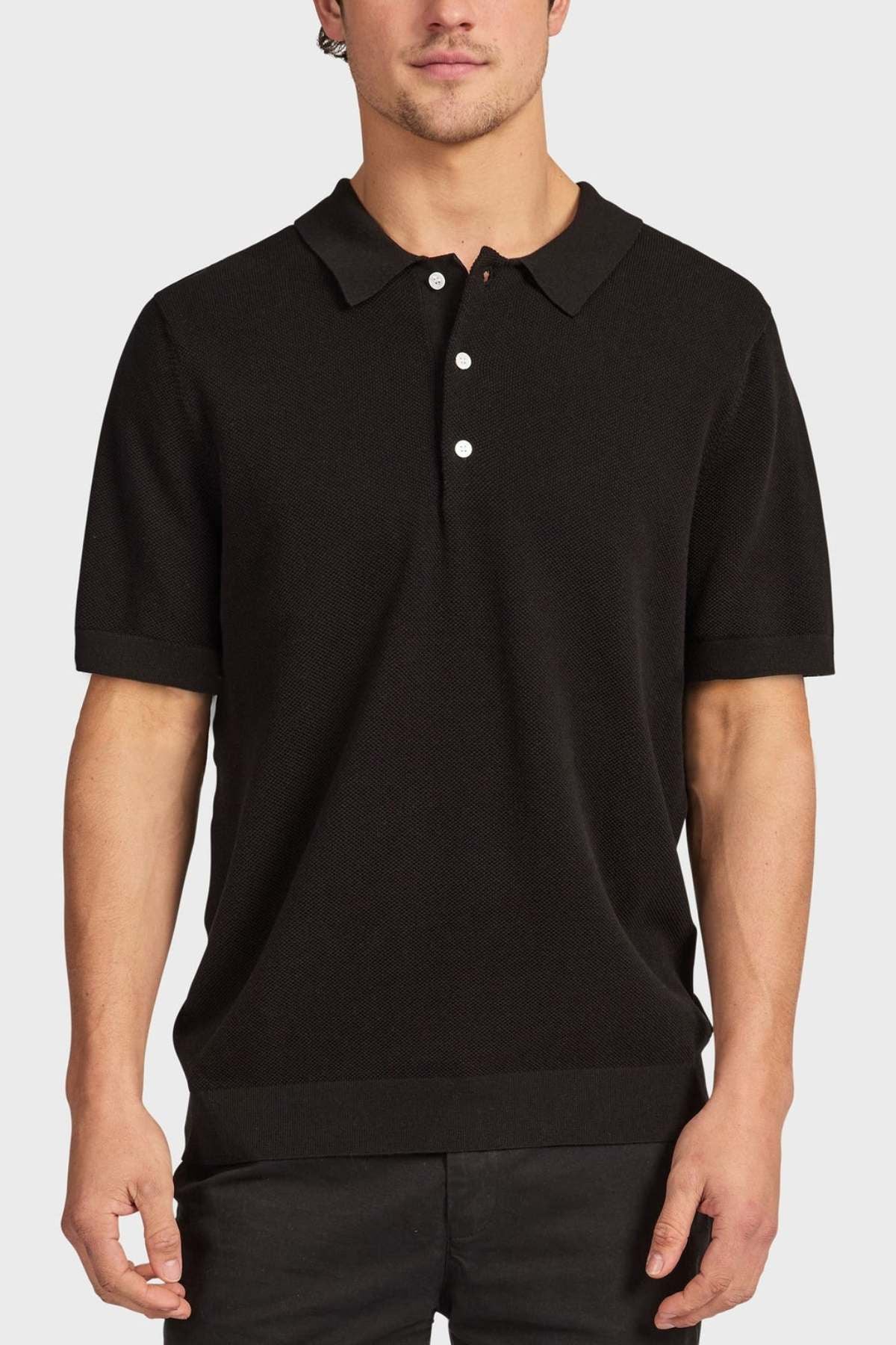 A man stands against a plain, light background wearing the Academy Brand Riley Knit Polo in black, featuring short sleeves, a collar, and three white buttons. The breathable fabric provides comfort as his arms rest relaxed by his sides.