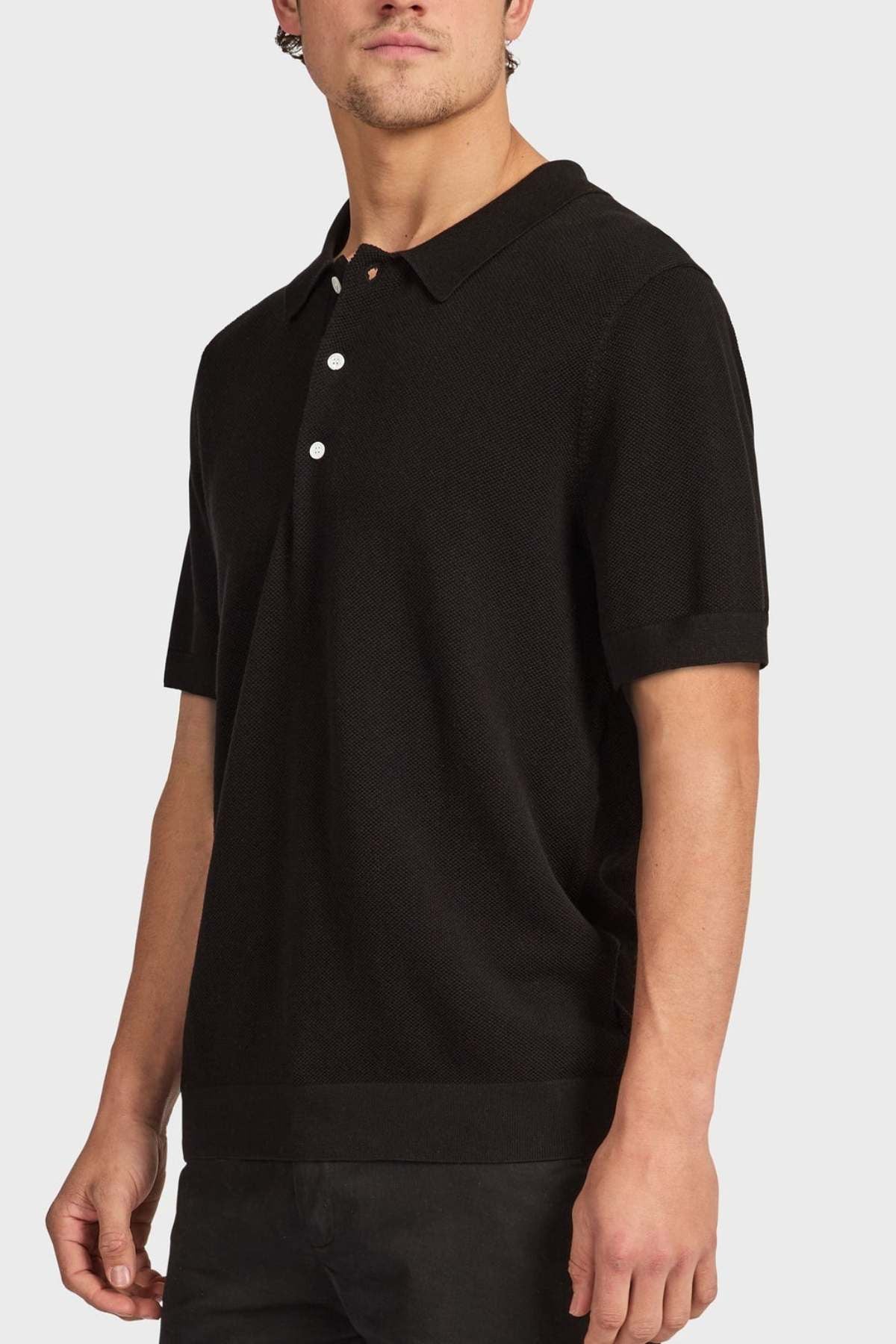 A man stands against a plain light background wearing the Academy Brand Riley Knit Polo in black, featuring three white buttons and crafted from breathable fabric.