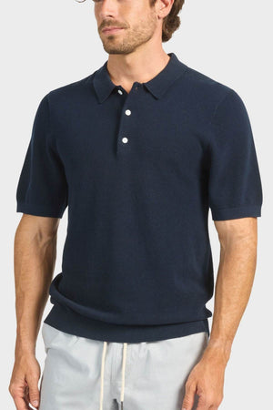 A man stands against a plain light background wearing the Academy Brand Riley Knit Polo in navy with white buttons and light, breathable drawstring pants.
