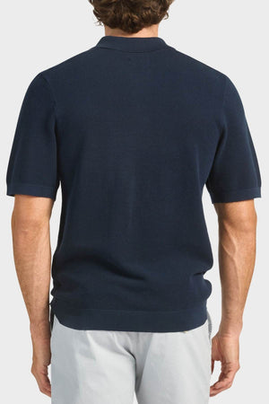 A man with short brown hair, seen from behind, is wearing a dark navy Academy Brand Riley Knit Polo made from breathable fabric and light gray pants, standing against a plain white background.