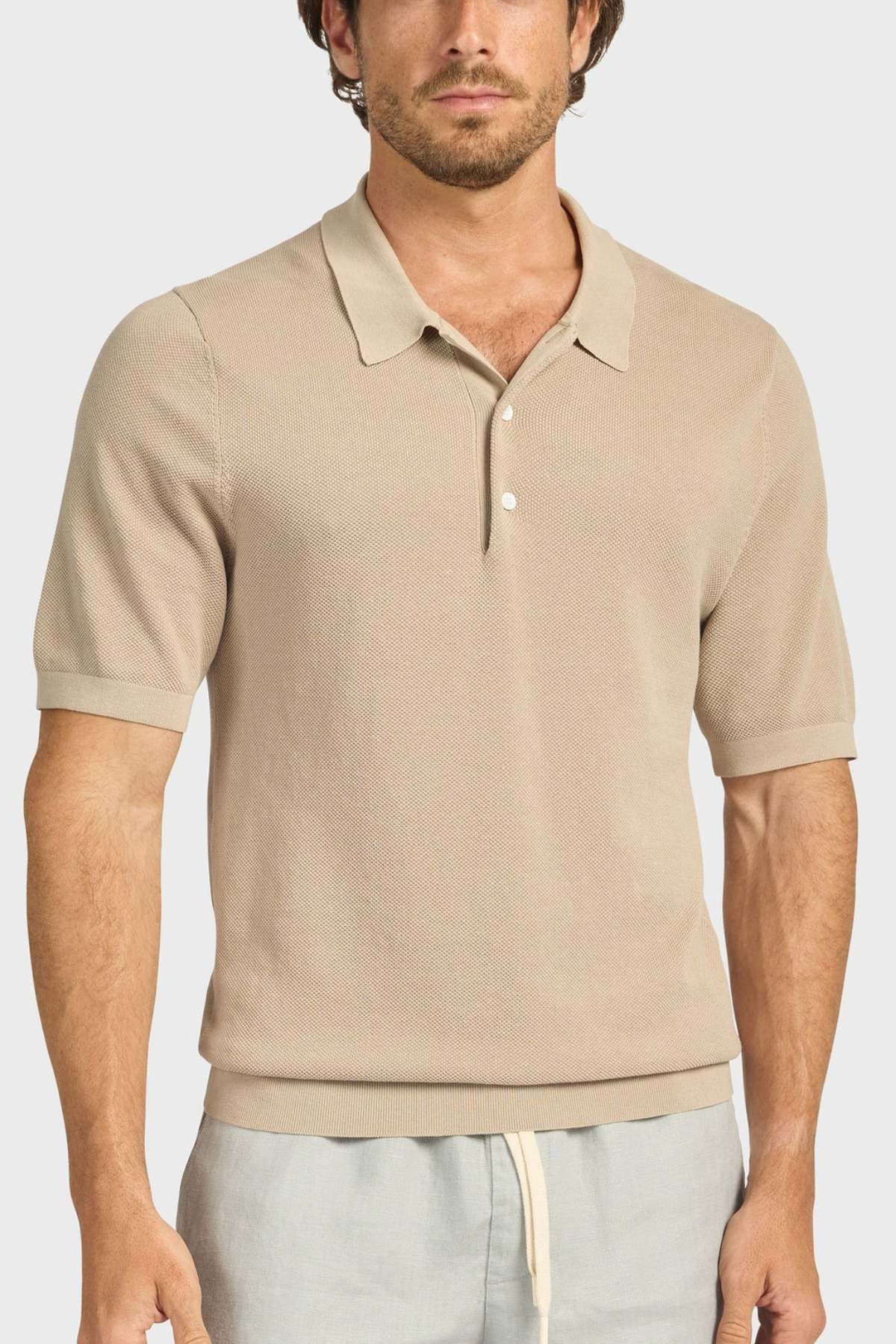 A man wears the Academy Brand Riley Knit Polo, a beige textured short-sleeve collared shirt with two buttons, paired with light gray drawstring pants, standing against a plain light background.