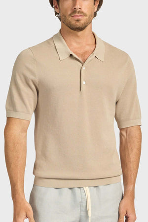 A man wears the Academy Brand Riley Knit Polo, a beige textured short-sleeve collared shirt with two buttons, paired with light gray drawstring pants, standing against a plain light background.