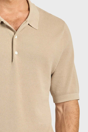 A close-up of a man wearing the Academy Brand Riley Knit Polo in beige, with textured stitching and two white buttons. The breathable fabric contrasts with the plain light background, highlighting his chest, arm, and collar.