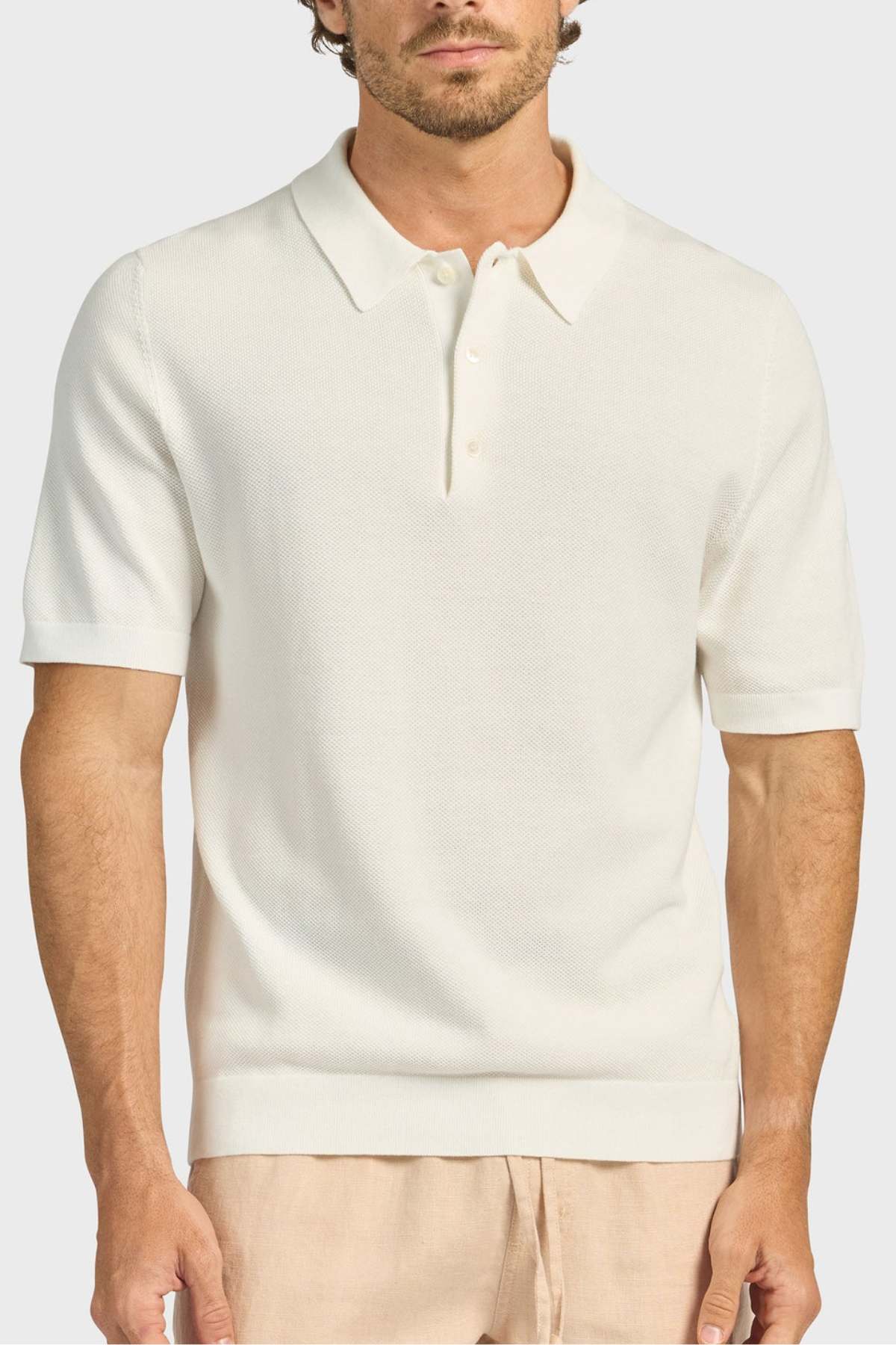 A man wears the Academy Brand Riley Knit Polo—short-sleeve, cream with a collar and three buttons—made from breathable fabric, paired with light beige pants, standing against a plain light background.