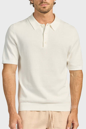 A man wears the Academy Brand Riley Knit Polo—short-sleeve, cream with a collar and three buttons—made from breathable fabric, paired with light beige pants, standing against a plain light background.
