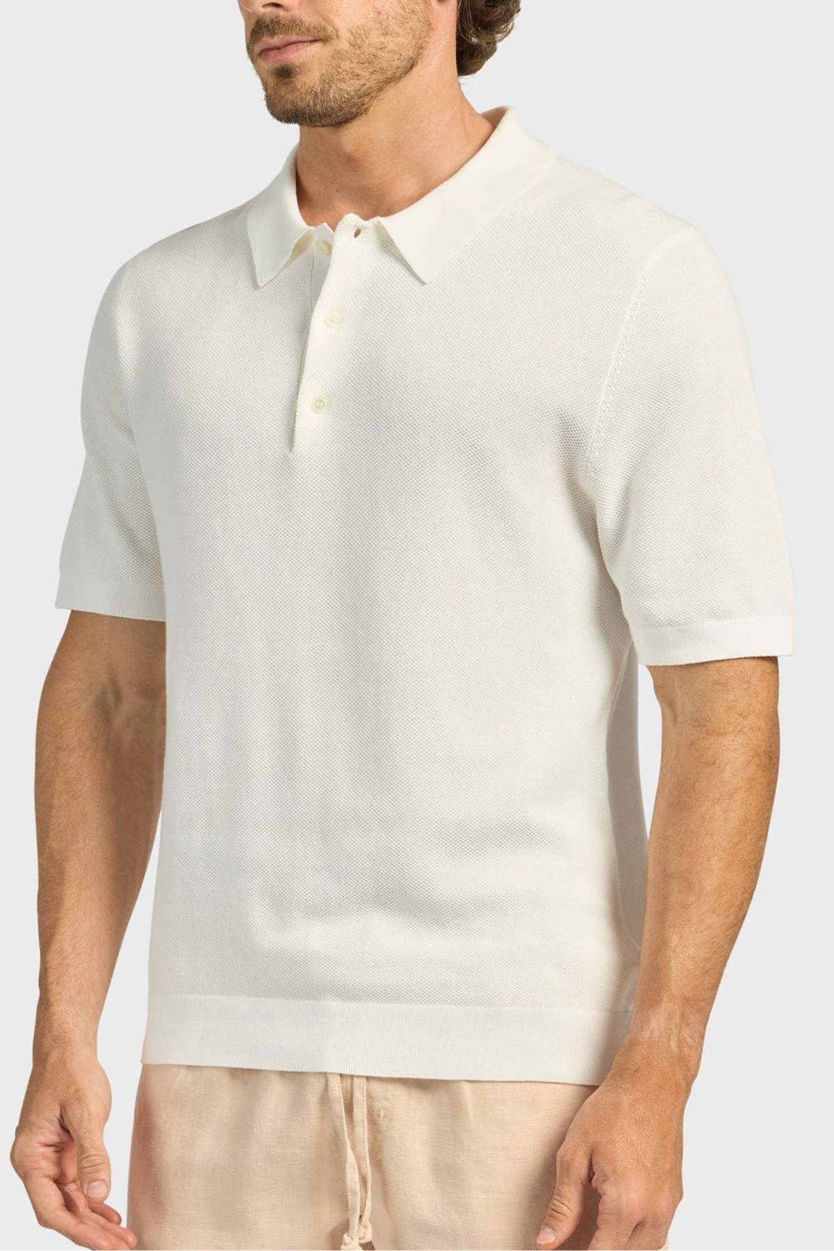 A man wears the Academy Brand Riley Knit Polo, a short-sleeve off-white textured polo made from breathable fabric, paired with beige pants, standing against a plain light gray background.