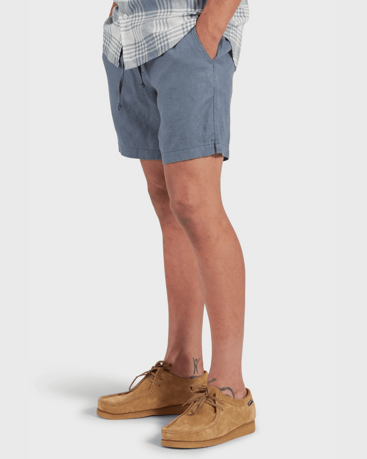 A person stands with hands in pockets, wearing the Academy Brand Riviera Linen Short in blue with an elastic waist, paired with a plaid shirt and tan suede shoes. Legs are visible, revealing a small tattoo above the right ankle. The background is plain and light.