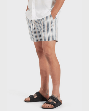 Wearing a white shirt, Academy Brand Teamster Stripe Boardy quick-dry recycled polyester shorts, and black buckle sandals, they stand against a plain white backdrop with hands in pockets, exuding a vintage vibe.