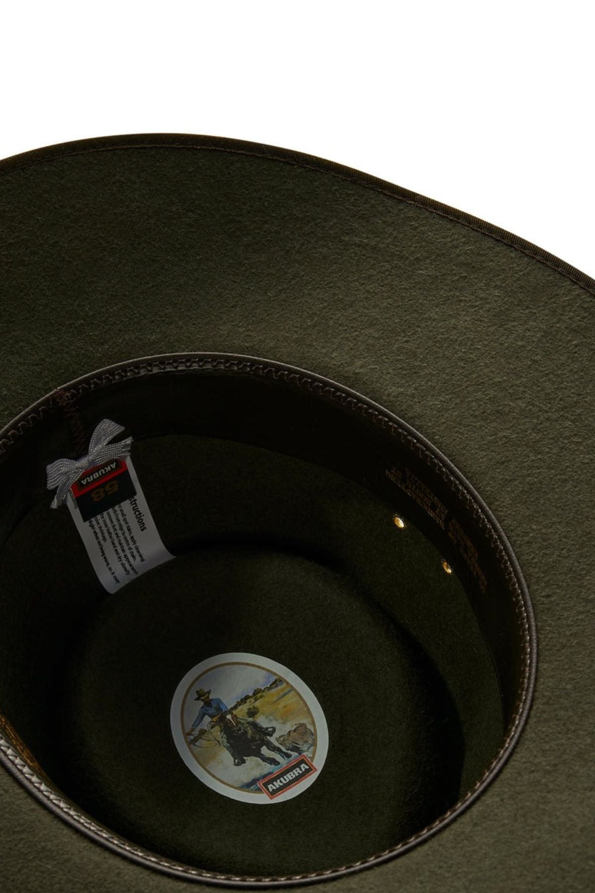 The image shows the interior of an Akubra Cattleman - Limited Edition cowboy hat in olive green. It features a woven label, a decorative sticker illustrating a cowboy on horseback, and intricate stitching along the brim, encapsulating Australian identity.
