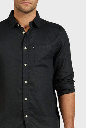 A man wears the Academy Brand Hampton Linen Shirt—a long-sleeved, dark linen shirt with a classic point collar, white buttons, and a chest pocket. His sleeves are rolled up, showing only the lower half of his face and upper body.
