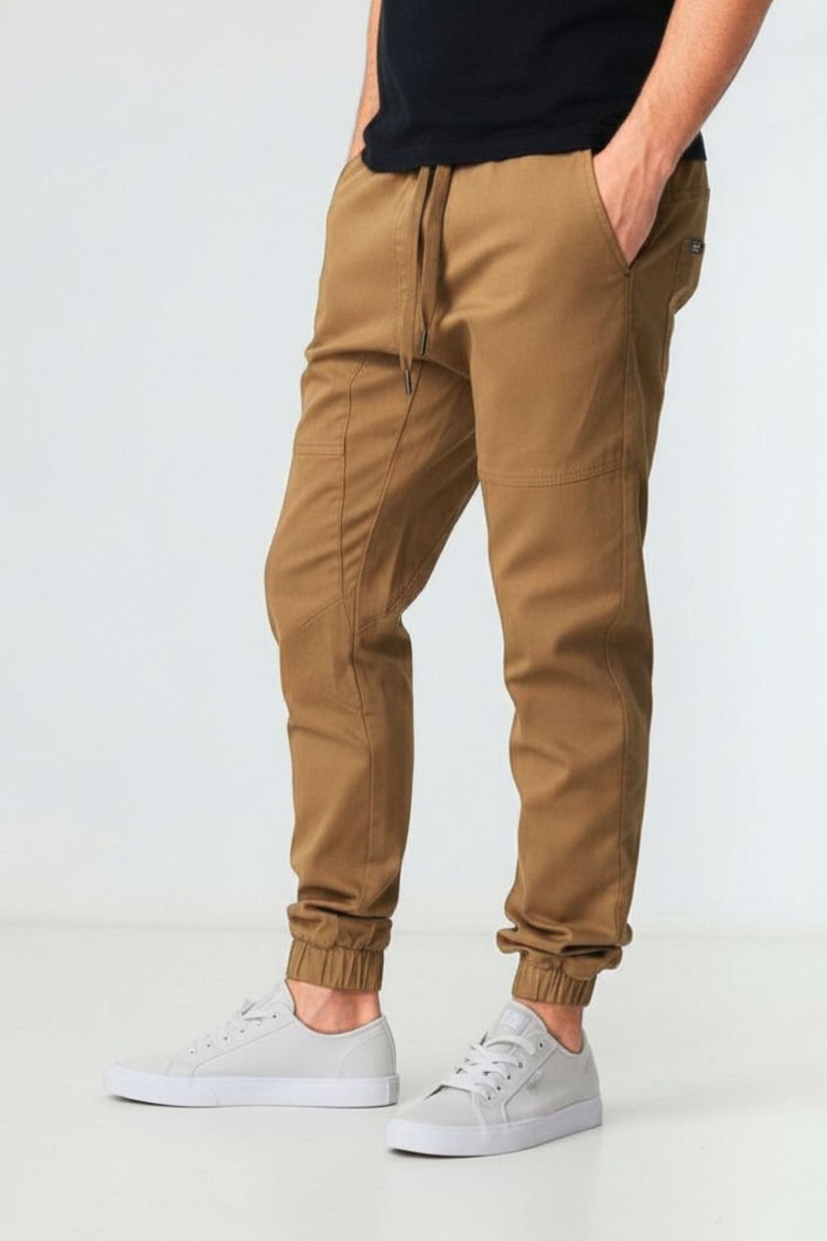 Wearing tan BLKWD James Jogger Pants made from knitted cotton stretch with elastic cuffs, a person stands with hands in pockets against a plain light background, paired with a black shirt and white sneakers.