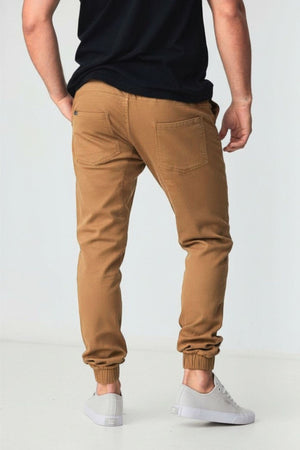 A man faces away, wearing BLKWD James Jogger Pants—men's joggers made from knitted cotton stretch with elastic cuffs—paired with a black t-shirt and white sneakers, standing against a plain light background.