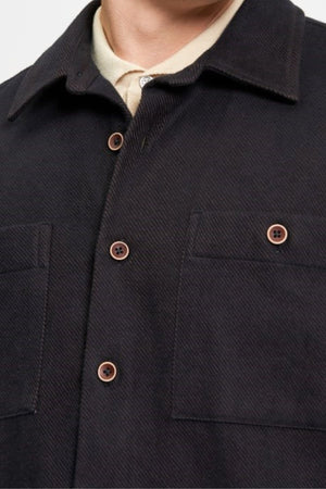 Close-up of a person's upper torso wearing the Ben Sherman Brushed Overshirt, a dark button-up with two chest pockets, brown buttons, and a pointed collar, layered over a light-colored collared shirt.