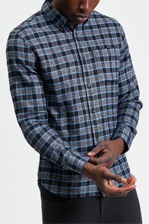 A man wearing the Ben Sherman Diffused Gingham Check Shirt in blue and black stands against a plain white background, adjusting his left sleeve with his right hand.