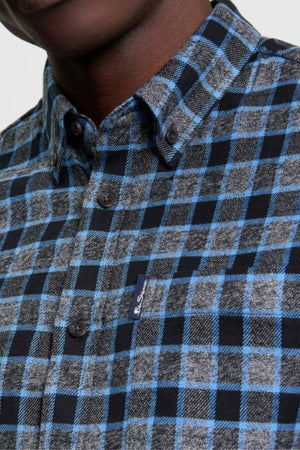 Close-up of a person wearing the Ben Sherman Diffused Gingham Check Shirt, featuring blue, black, and gray plaid with a front pocket and visible small fabric tag; part of the neck and lower face is shown.