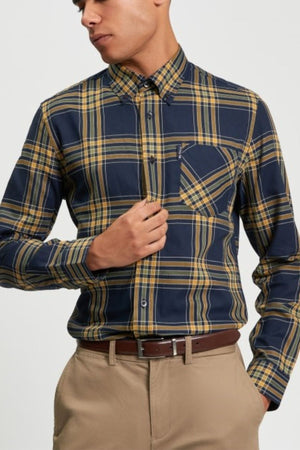 A man wearing the Ben Sherman Grid Check Shirt, navy blue and yellow plaid with a front pocket, pairs it with tan pants and a brown belt. He stands with his head slightly turned, one hand adjusting his shirt.