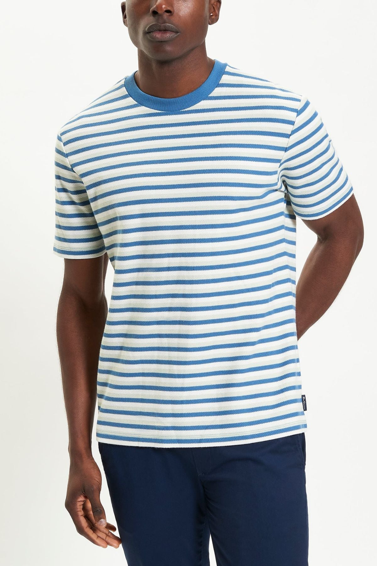 A person stands against a plain white background wearing the Ben Sherman Honeycomb Stripe Tee—short-sleeved with white and blue horizontal stripes and a blue collar—paired with dark blue pants.