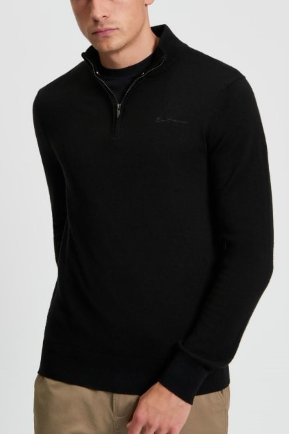 A man wears the Ben Sherman Merino Half Zip Knit in black over a black shirt and beige pants, standing with one hand in his pocket against a plain background.