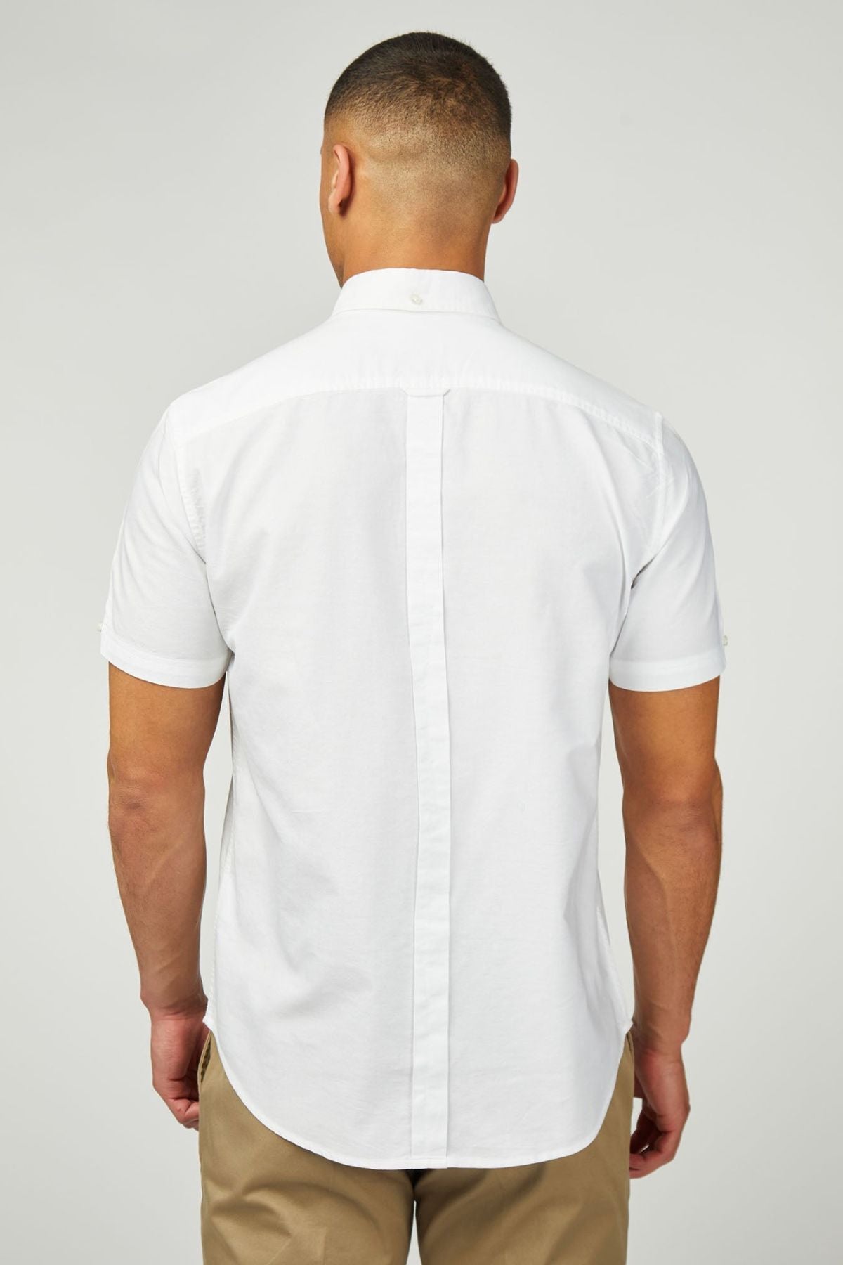 A man stands with his back to the camera, wearing a white Ben Sherman Signature Oxford Short Sleeve Shirt and tan pants, set against a plain light gray background.