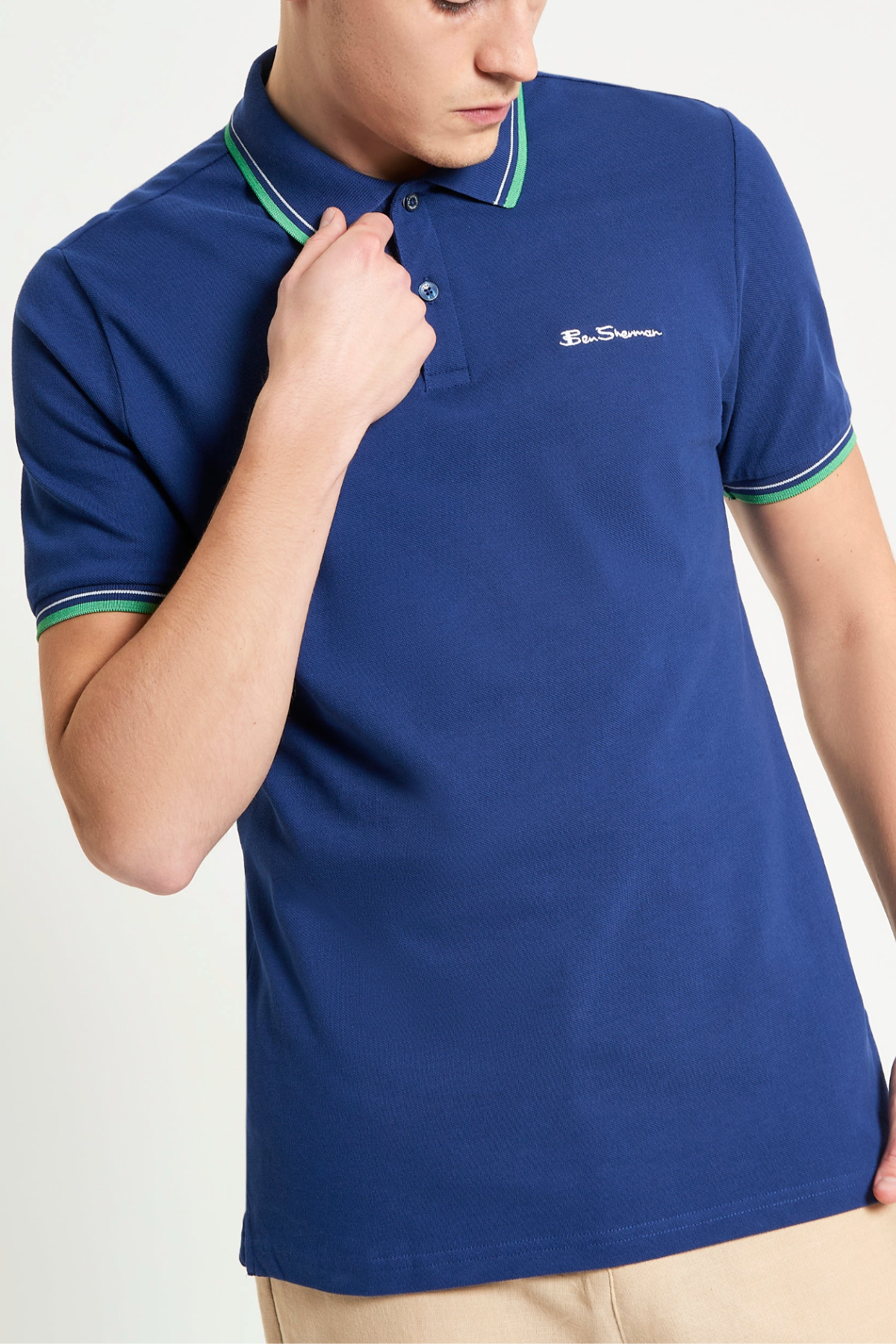 A person wears a Ben Sherman Signature Pique Polo in blue with green and white trim on the collar and sleeves, paired with beige pants. The shirt has a small embroidered logo on the left chest. The person’s head is partially out of frame.