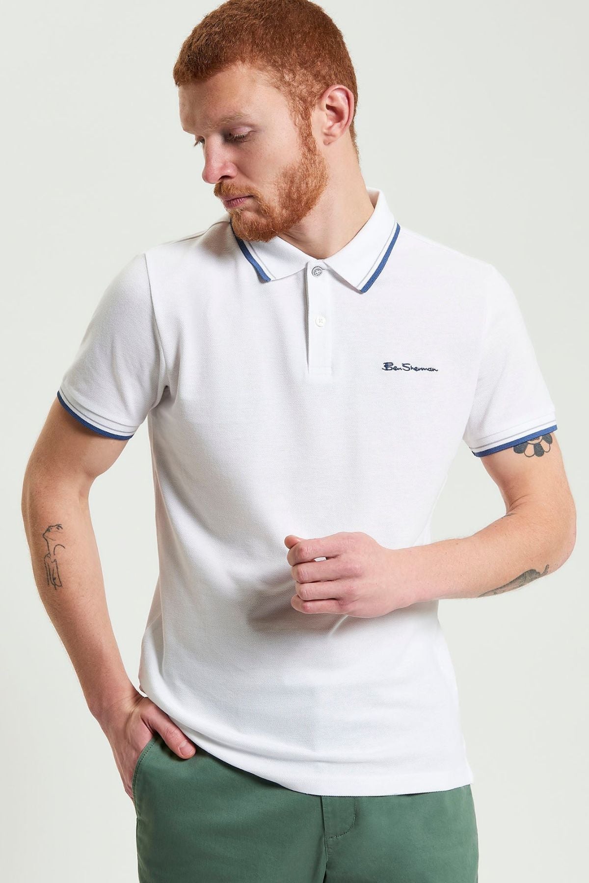 A man with short red hair and a beard wears a white Ben Sherman Signature Pique Polo with blue trim and green pants. Made from organic cotton pique, he stands with one hand in his pocket, looking down to his left.