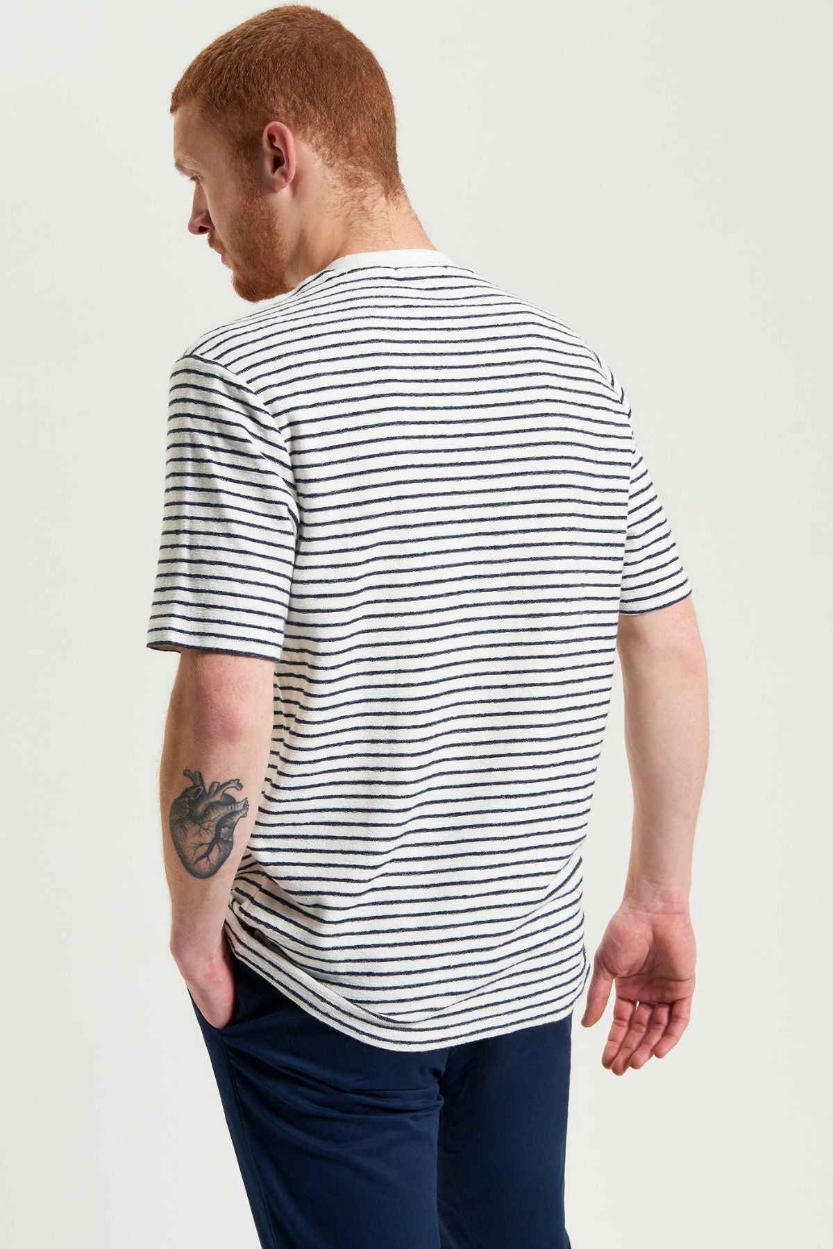 A man with short red hair and a beard, wearing a Ben Sherman Textured Stripe Tee and navy pants, stands with his back to the camera. A heart-shaped tattoo is visible on his right forearm.