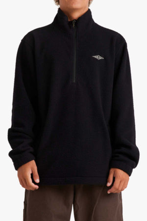 Wearing the Billabong A/Div Boundary Mock Neck Sweatshirt, a black fleece pullover made of recycled polyester with a half-zip and an embroidered chest logo, a person stands relaxed in brown pants. The image is cropped at the neck and mid-thigh.