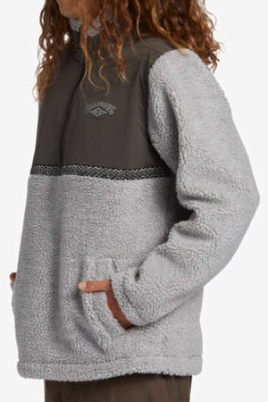 Wearing the Billabong A/DIV Boundary Re-Issue Sweatshirt—a fuzzy light gray sherpa pullover with a dark gray chest panel and zip collar—this long-haired individual shows off a cozy, eco-friendly staple for outdoor adventures.