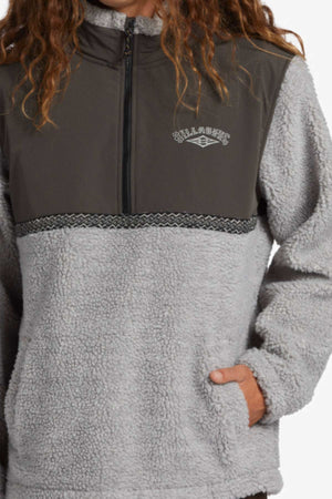 A person with long, wavy brown hair wears the Billabong A/DIV Boundary Re-Issue Sweatshirt—a gray, fuzzy, recycled polyester half-zip with side pockets, a darker gray chest panel, and "Billabong" logo on the left chest. Only upper body is visible.