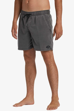 A barefoot person against a white background wears gray Billabong All Day Overdye Layback Shorts featuring an elastic waistband, side pockets, and a relaxed fit. Made from eco-conscious fabric, the shorts also display a small patch logo on the left leg.