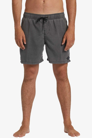 A person barefoot against a white background wears Billabong All Day Overdye Layback Shorts, crafted from eco-conscious fabric. The gray shorts have two front pockets and a small logo on the left leg, and the person's hands rest naturally at their sides.