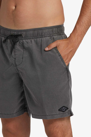 A person models Billabong All Day Overdye Layback Shorts in gray, made from eco-conscious fabric, with one hand in a pocket. The shorts show a black logo patch near the hem, and the scene is shot waist-down against a plain background.