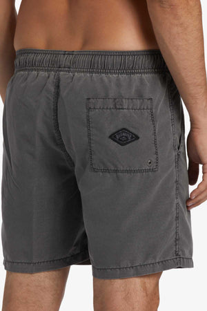 A person in Billabong All Day Overdye Layback Short wears dark gray swim trunks with an elastic waistband crafted from eco-friendly fabric, featuring a visible back pocket and a diamond-shaped logo patch on the lower back.