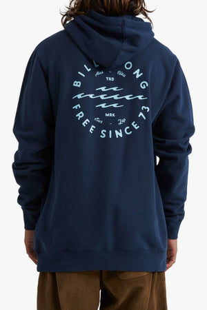 A person with long hair is seen from the back wearing a dark blue Billabong Big Wave Daz Pop Hood, perfect for casual outings. This comfortable half brushed fleece hoodie features a circular graphic with wavy lines and the text "Billabong Free Since '73" printed in white on the back. The person is also wearing brown pants.