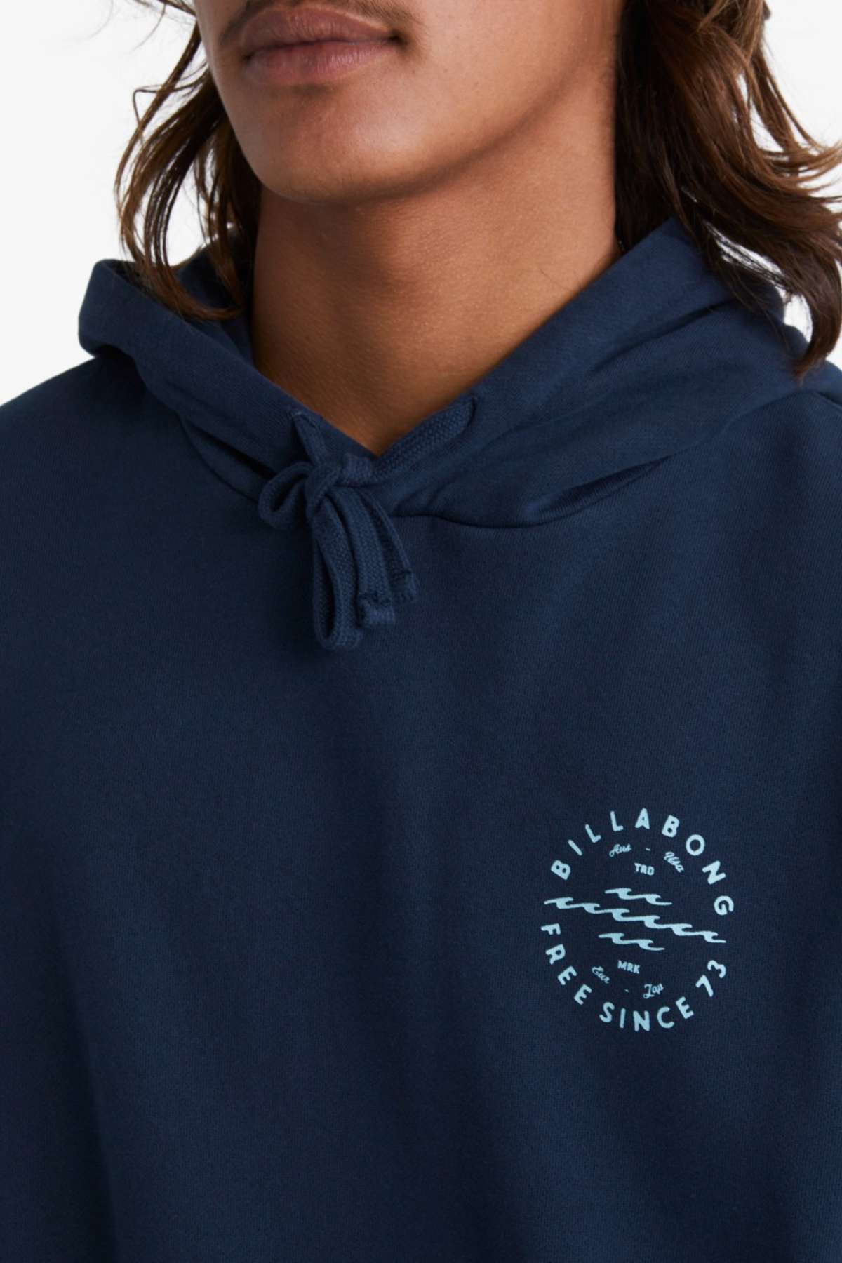 A person with long hair is seen wearing a navy Billabong Big Wave Daz Pop Hood hoodie. This comfortable piece, crafted from half brushed fleece, showcases a round printed design on the chest that reads "Billabong Free Since '73" alongside a stylized wave graphic. Ideal for casual outings, the hoodie is finished with drawstring ties at the neck.