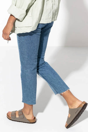 A person is shown from the waist down wearing a light green shirt, blue cropped jeans, and taupe Birkenstock Arizona sandals with adjustable buckles and a contoured footbed, walking on a white background.