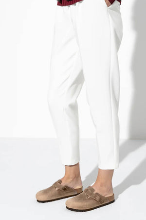 A person wearing white cropped pants and light brown Birkenstock Boston clogs with an anatomically shaped footbed stands against a white background.