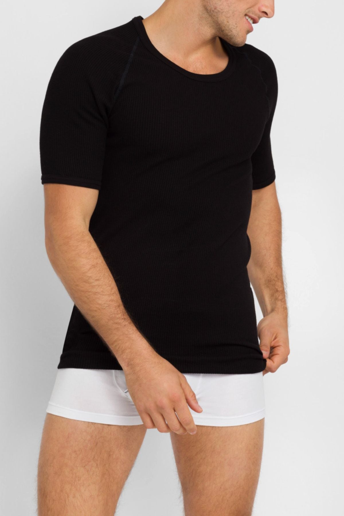 A man wearing a Bonds Aircel Short Sleeve Tee in black and white boxer briefs stands against a plain light background, looking to his right while slightly pulling down the hem of his shirt.