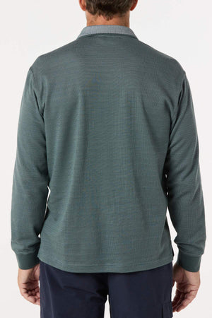 A man is seen from behind wearing a Breakaway Avantee Micro Prima Polo in textured teal with long sleeves, rib knit cuffs, and navy pants. Set against a plain white background, the look combines classic charm with modern style.