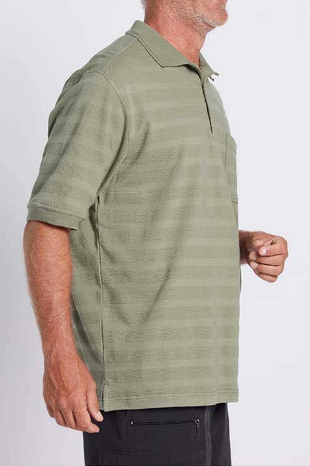A person with short hair, dressed in a green Breakaway Emil Polo with self-jacquard stripes and black pants. Their face is turned slightly to the side, making it not visible, and they seem to be standing against a plain background.