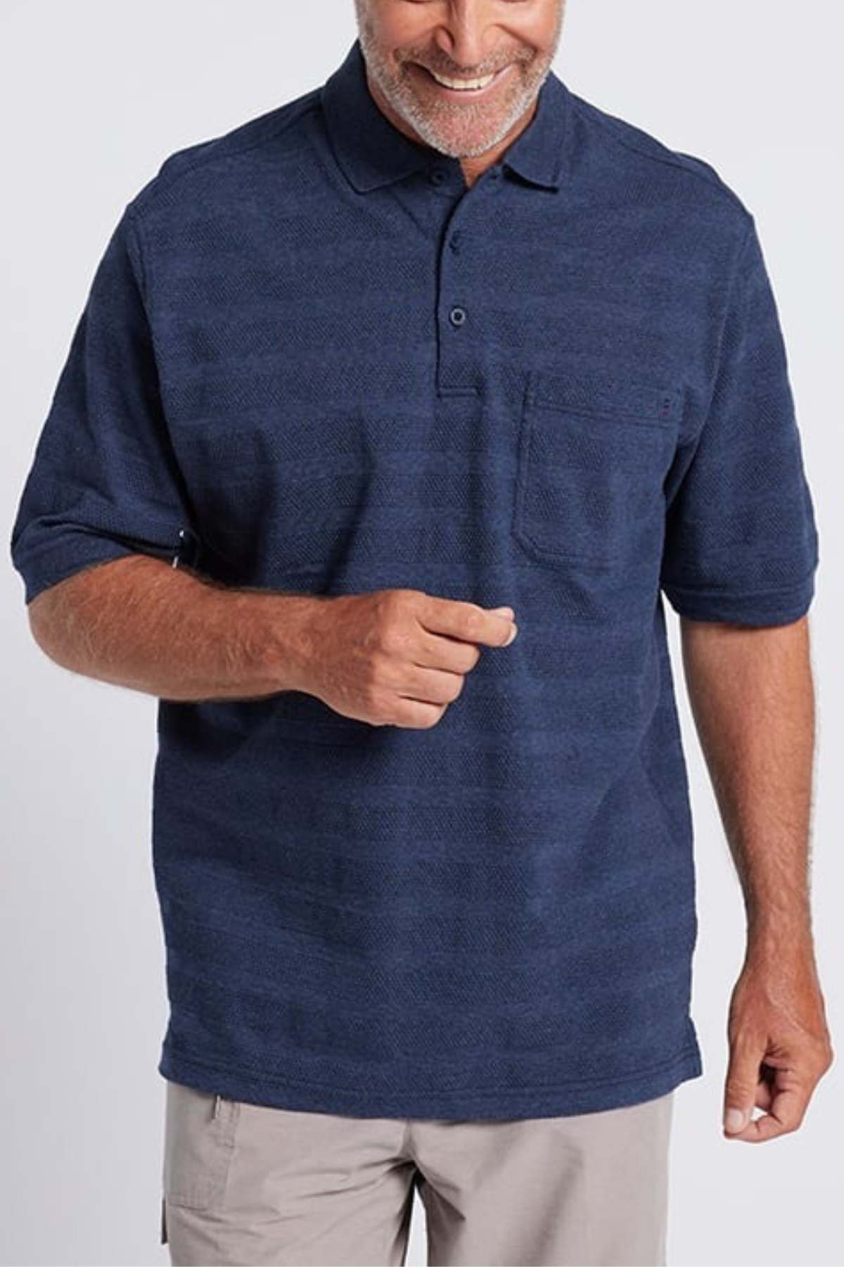 A man with a short beard and a friendly smile is wearing the Breakaway Emil Polo, which has an elegant knit collar and is made from 100% cotton. The blue polo shirt includes a chest pocket, and he pairs it with khaki pants. His right hand is positioned in front of his body while his left arm rests relaxed by his side against the white background.