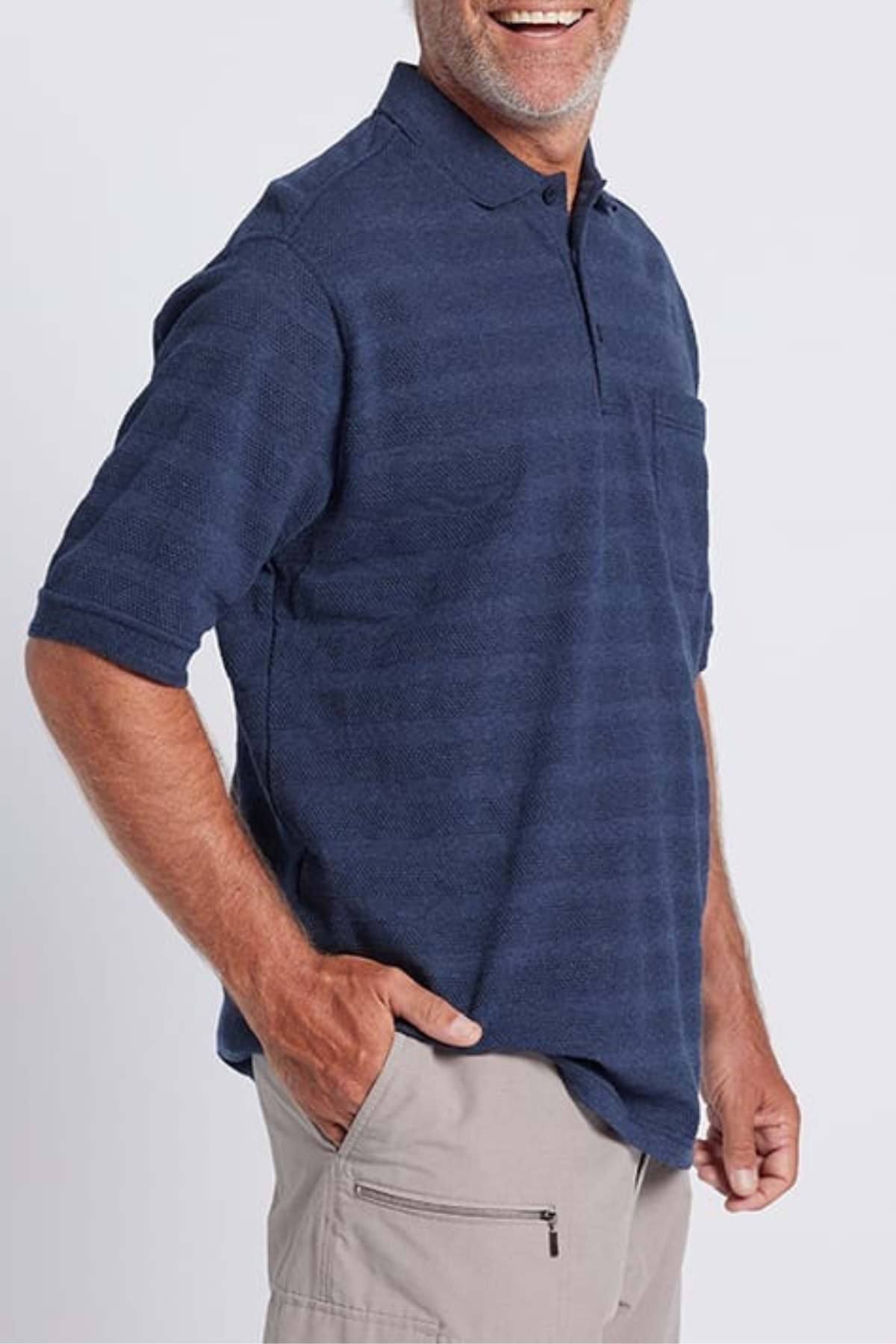 A person wearing a navy blue Breakaway Emil Polo with an elegant knit collar and light gray shorts with a zipper pocket stands against a plain light background. The 100% cotton polo looks great as they have one hand in their pocket and are smiling.