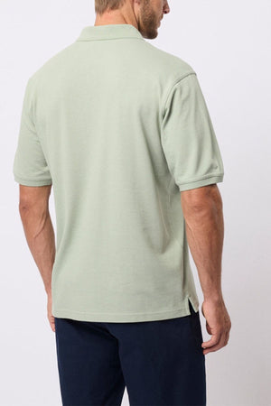 A man stands with his back to the camera, wearing a light green Breakaway Fraser Cotton Pique Polo and dark navy pants. The polo's color stands out against the plain, light background.