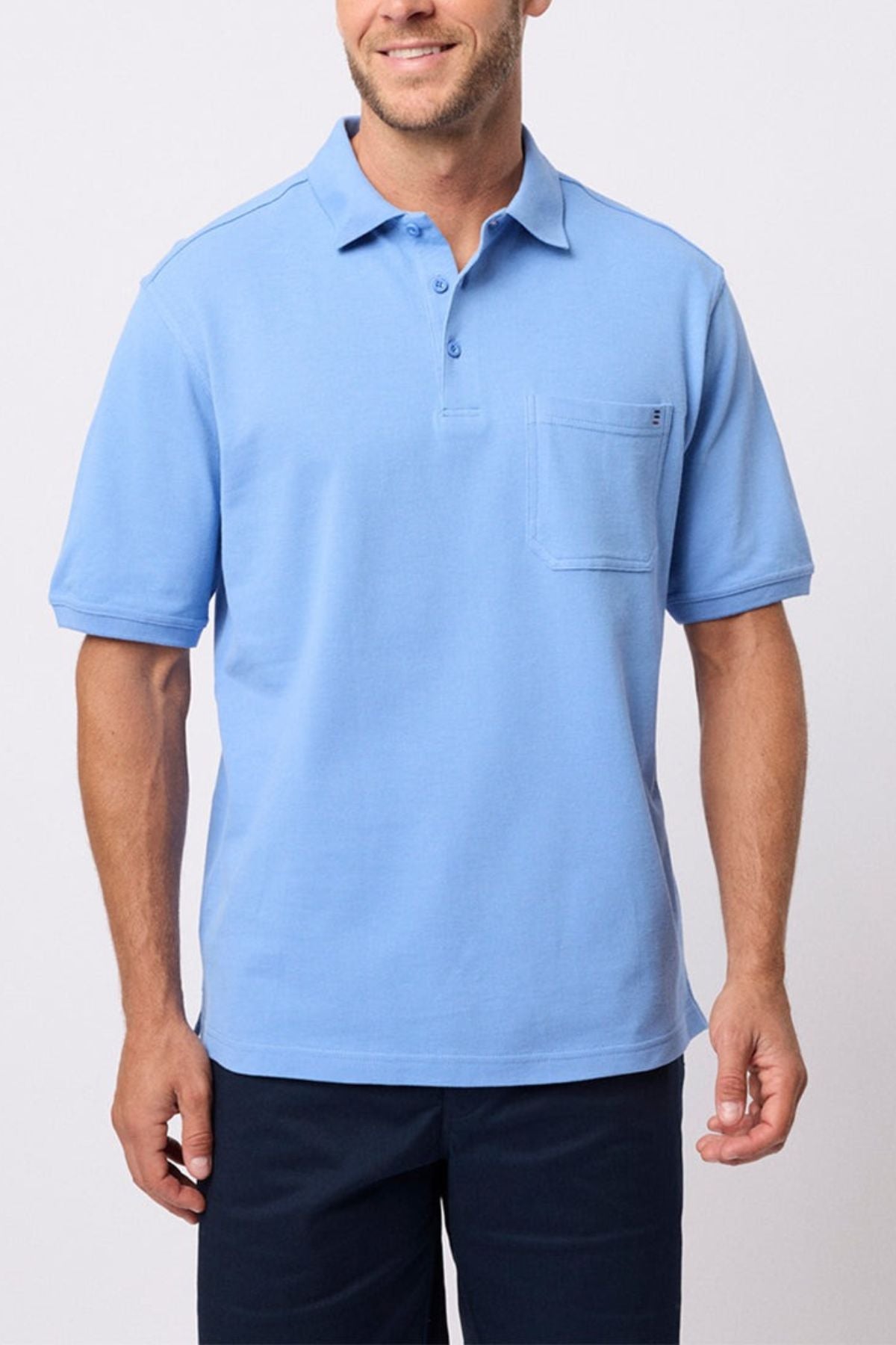 A man stands against a plain light background wearing the Breakaway Fraser Cotton Pique Polo in light blue, featuring soft cotton pique fabric and a chest pocket, paired with dark pants.