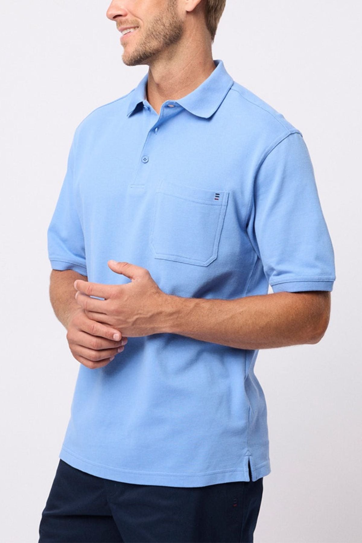 A man wearing the Breakaway Fraser Cotton Pique Polo in light blue with a chest pocket and dark shorts stands against a plain light background, smiling and looking to the side.