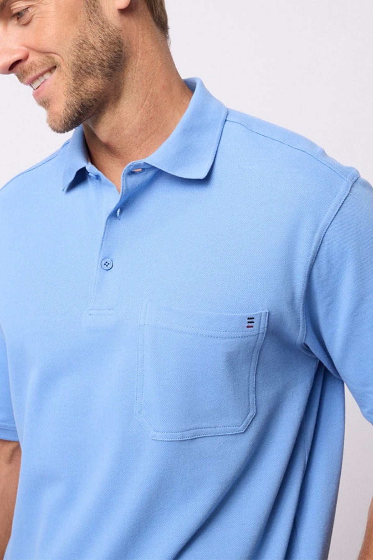 A man stands against a plain light background, smiling slightly and facing left, wearing the Breakaway Fraser Cotton Pique Polo in light blue with a front pocket.