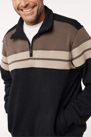 A man smiles with his hands in his pockets, wearing a Breakaway Henrich Snowy Mt Fleece Half Zip with wide black and brown stripes and beige accents over a white undershirt. Only his upper body is visible.
