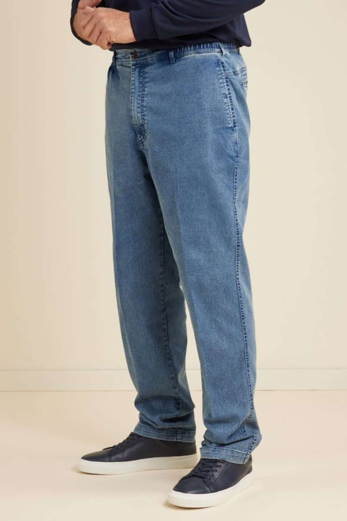 A person stands indoors wearing Breakaway Intense Denim Jeans with a straight leg design, paired with a dark long-sleeve shirt and navy blue sneakers; their hands are clasped in front and their face is not visible.